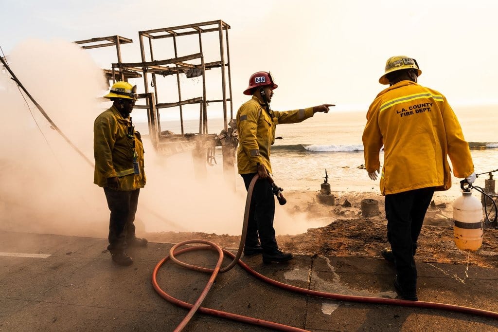 Three firefighters stand near the edge of what used to be a home overlooking the ocean that was burned during a wildfire. Part of a remaining structure can be seen in the background as smoke rises below the rubble.