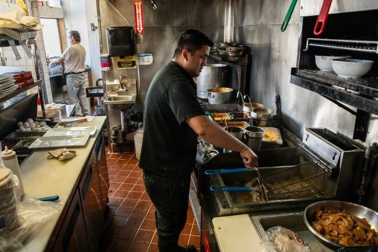 A cook dressed in a black outfit works in the kitchen of a restaurant, at the fryer. Another person, in the background, wearing denim and a white shirt, washes dishes.