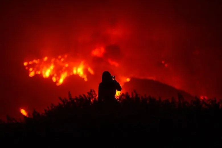 The silhouette of a person set against the background of a hill burning during a wildfire as they take a photo or video with their phone.