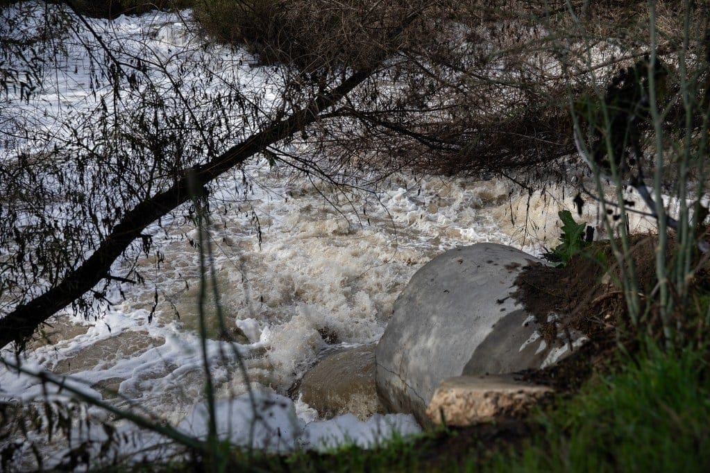 Layers of white foam caused by sewage and chemicals bubble up along a section of a river surrounded by greenery.