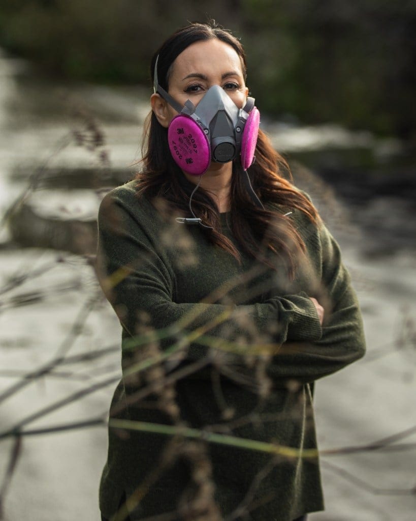 A person, wearing a pink respiratory filter mask and with their arms crossed, stands near a flooded area.